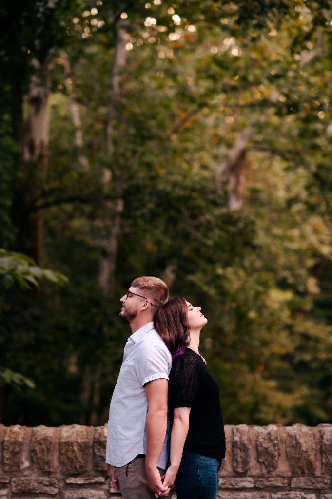 A couple stands back to back while holding hands and leaning into one another in front of a stone wall. 