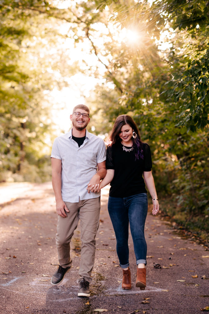 A couple walks together along a trail holding hands and smiling while the sun shines behind them. 