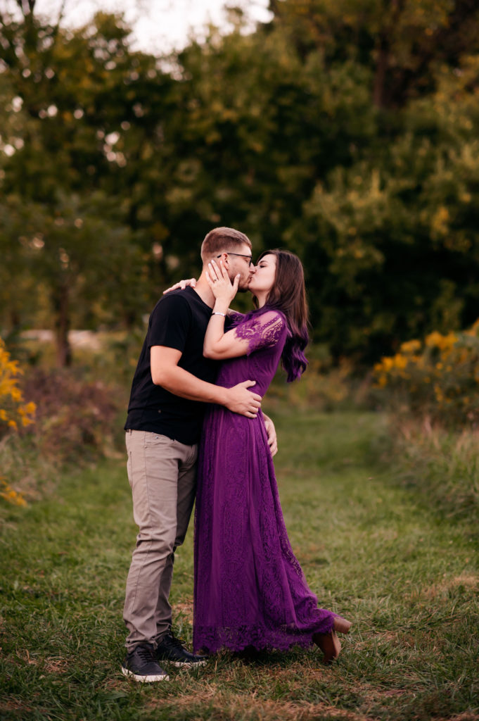 A couple stops for a kiss along a grassed trail through a field of tall grass. 