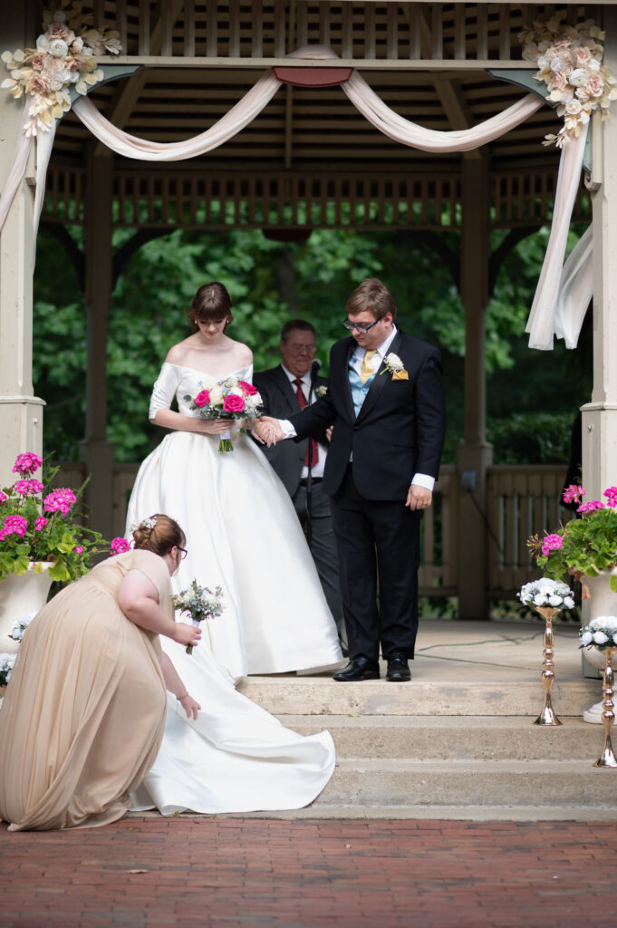 The maid of honor fixes the bride's train of her dress for her, while the couple holds hands waiting for their ceremony to begin. 