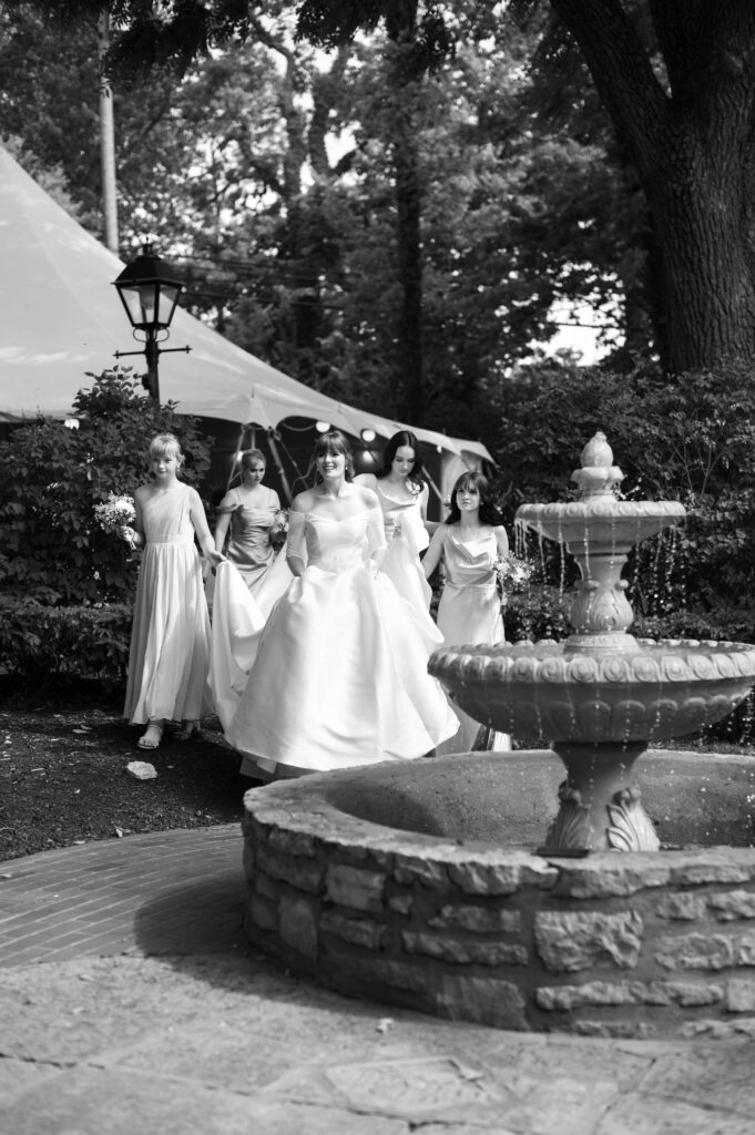 A black and white photo of a bride in her elegant wedding gown walking to the ceremony on her big day. She smiles big as bridesmaids help to carry her dress. 