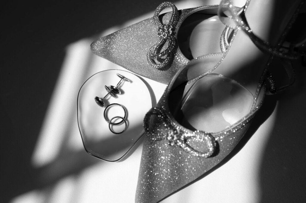 A black and white photo shows a deep shadow casted across wedding shoes, rings, and cufflinks, creating a moody yet elegant feel to the wedding photo. 