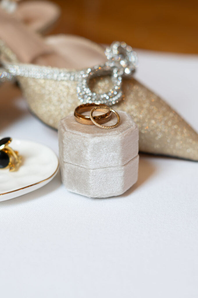 Two wedding bands sit atop a ring box, as they are the focus in the wedding photo, with blurred elegant details behind them. 